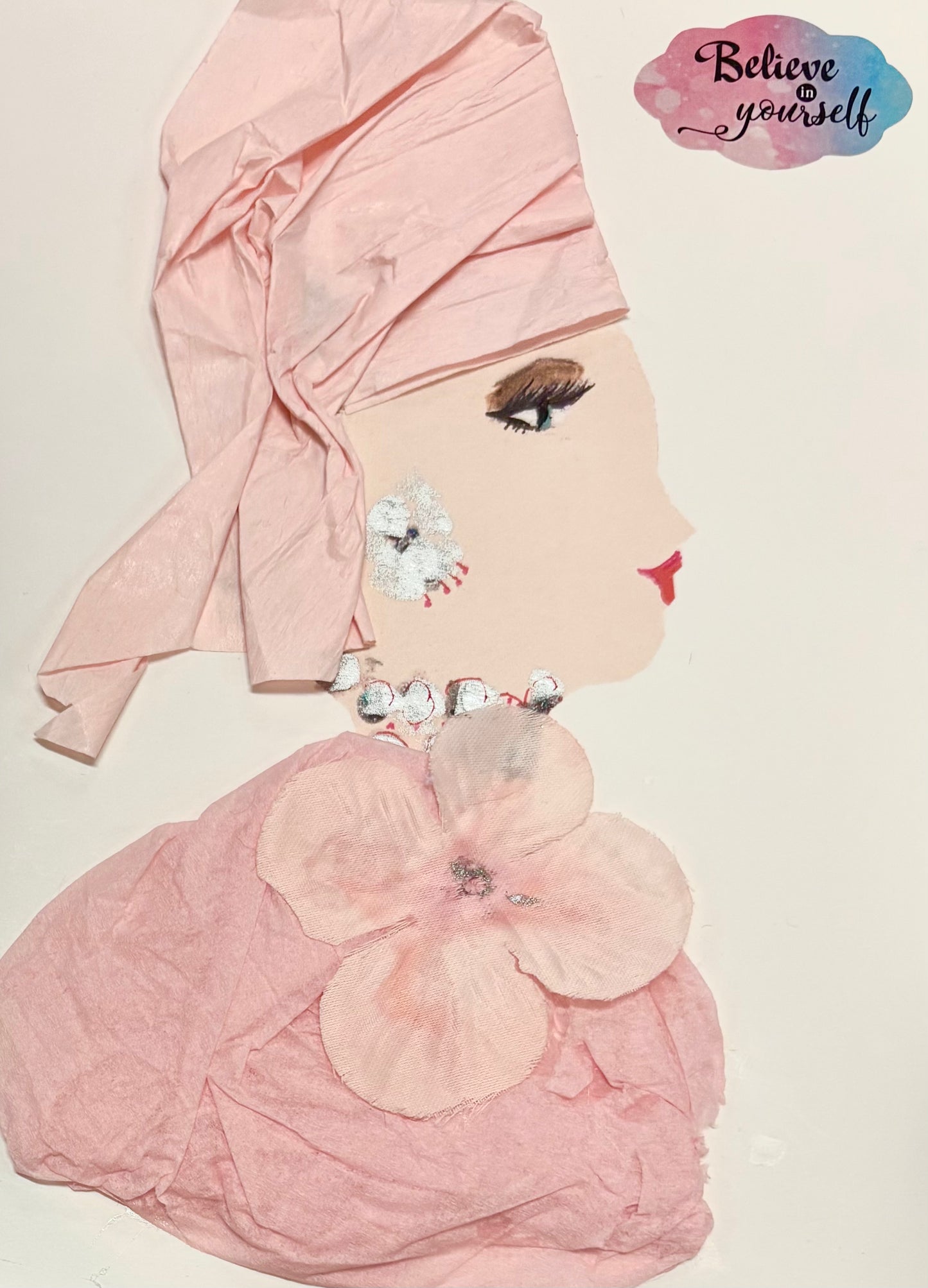 A woman wears a baby pink headwrap and blouse. The blouse has a baby pink flower on top. She wears silver jewellery. A sticker in the top right corner says "believe in yourself."
