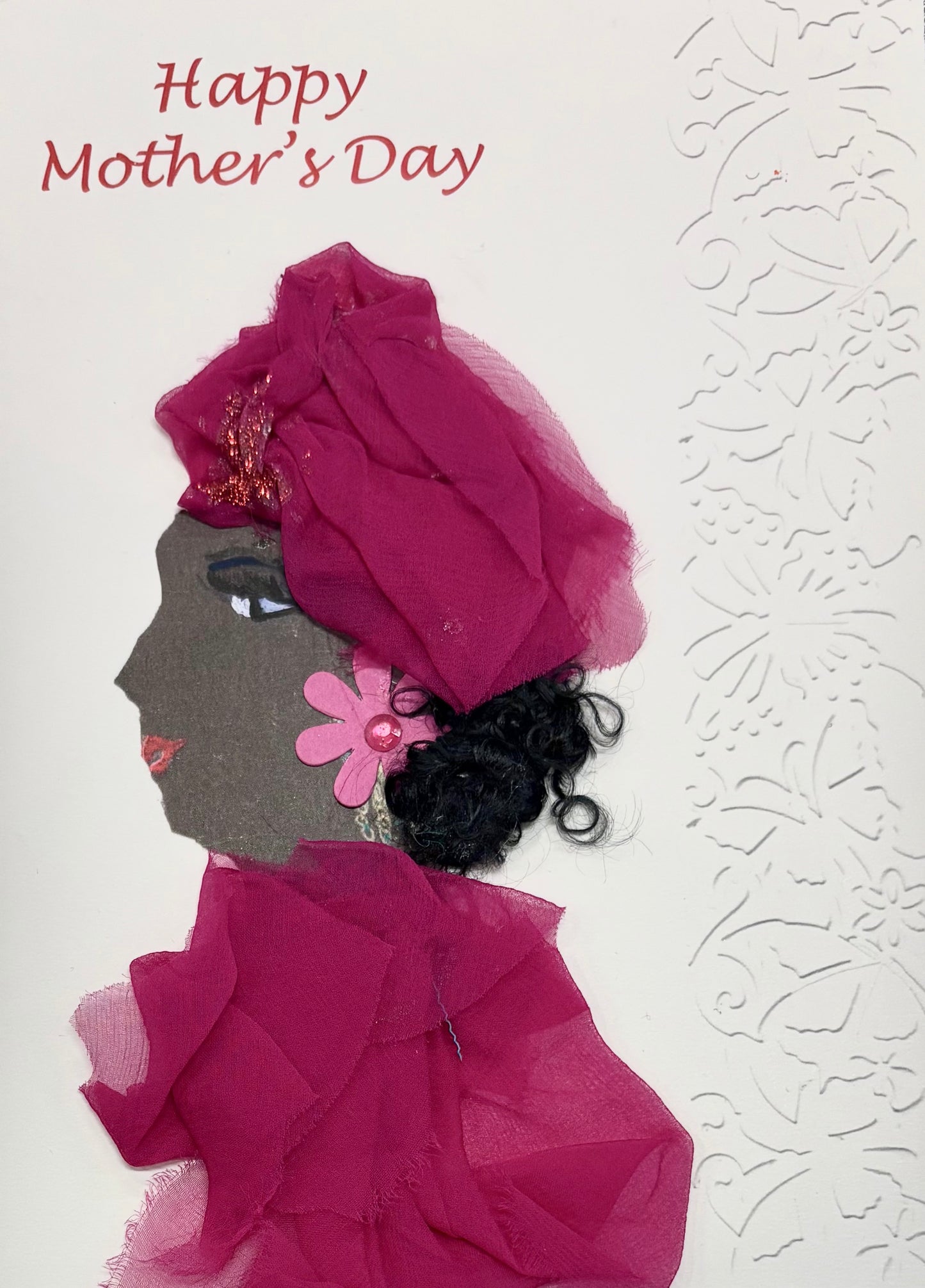 A woman wears a deep pink headwrap and blouse. She has a light pink flower earring. The right side of the card is textured with a subtle butterfly pattern. Happy Mother's Day is spelt out in the top left corner in red. 