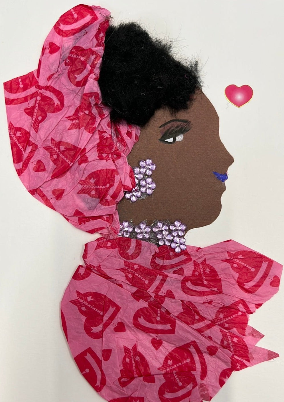 This card has been given the name Tilly Terrific. Tilly wears a matching headdress and blouse which are made of a recycled paper material. The pattern on the pink paper is a darker pink heart with an arrow going through it, then surrounded by smaller hearts. Her jewellery is made of small purple flower gems. To the right of her, there is a small pink heart.